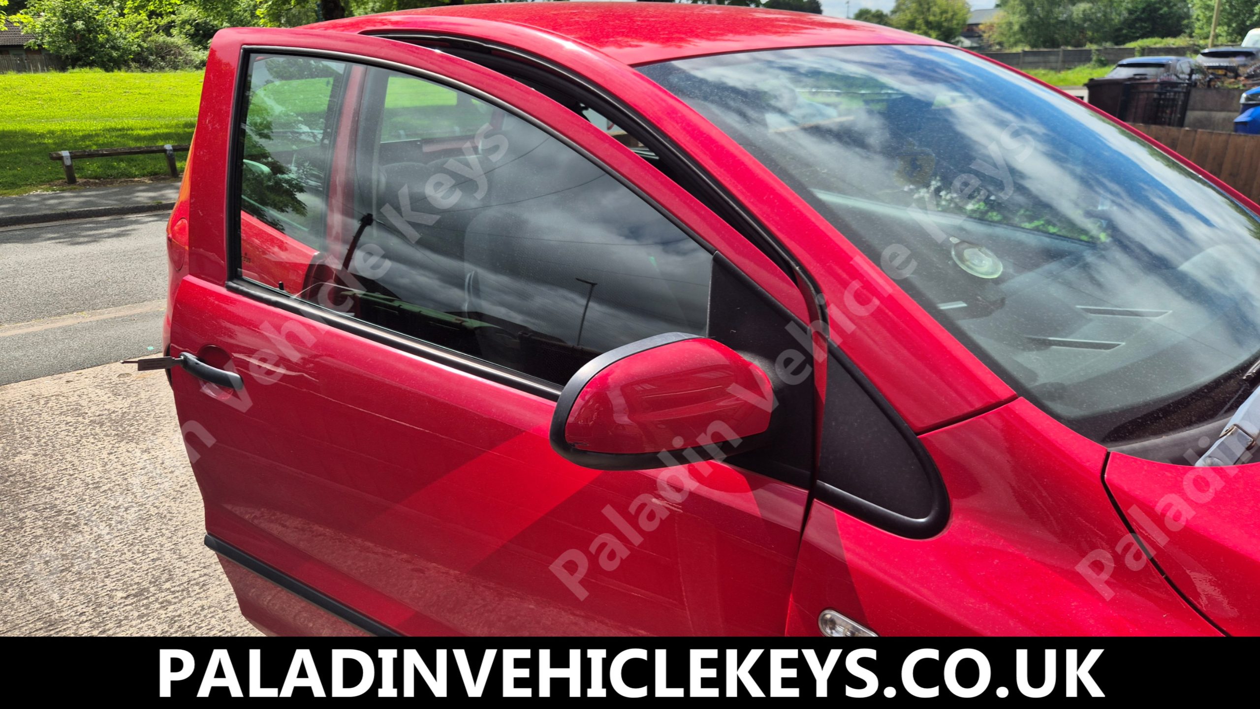 Close-up of a red car with the driver’s door open, showing the side mirror and window, parked on a street.